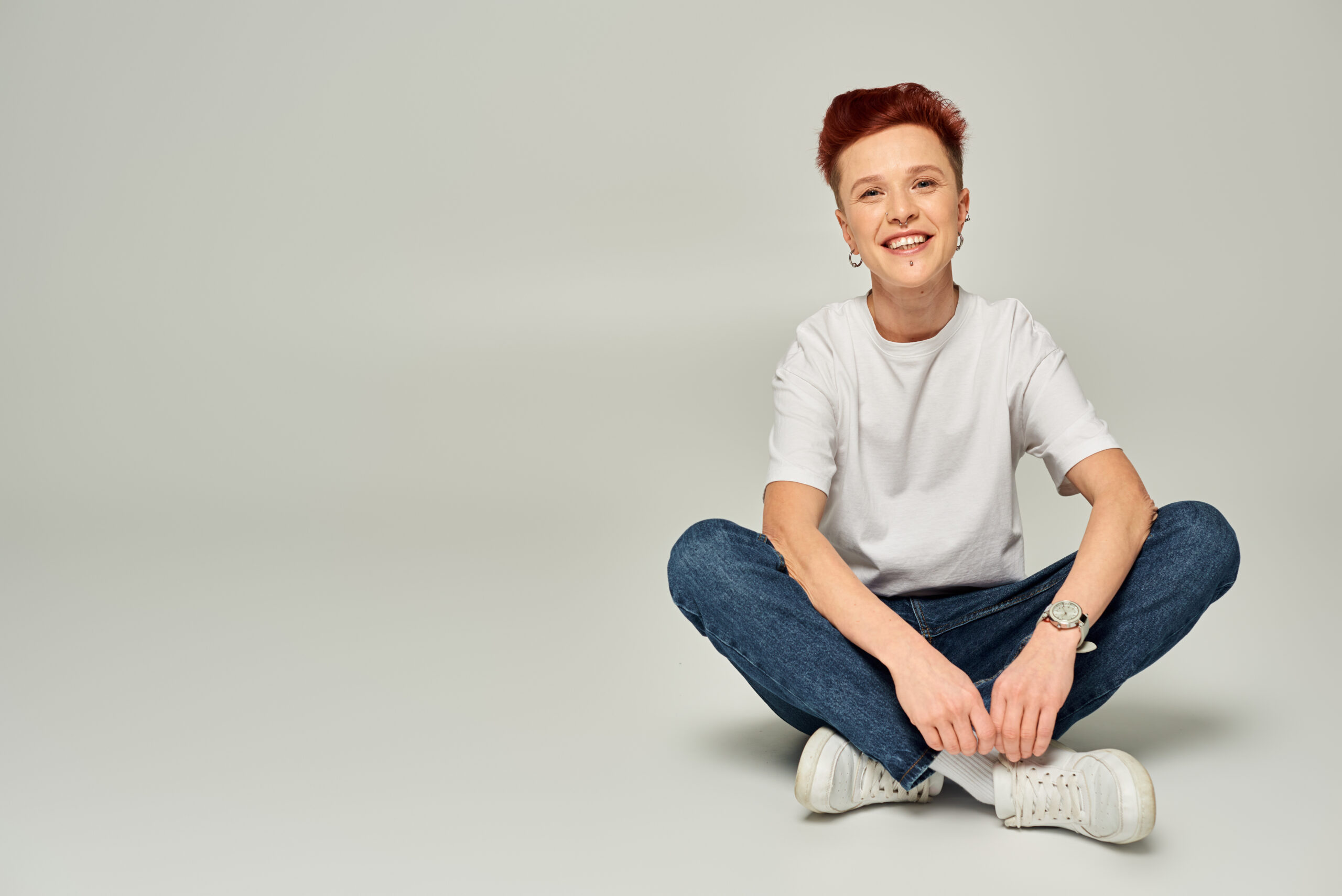 cheerful redhead non-binary person in white t-shirt and jeans sitting and looking at camera on grey non-binary person in white t-shirt and jeans sitting